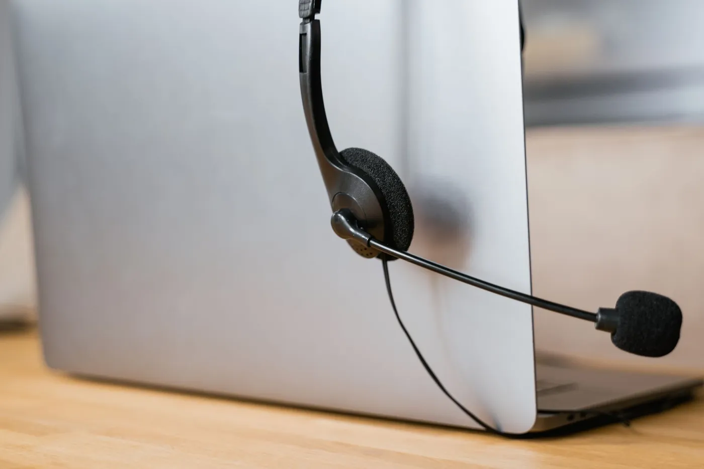 A support headset with a microphone hanging on the edge of an open laptop on a wooden desk